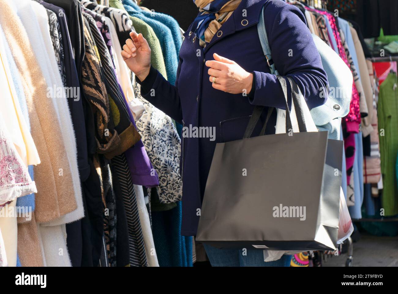 A woman shopping at an outdoor market, observes clothes hanging on racks and hangers in a city square Stock Photo