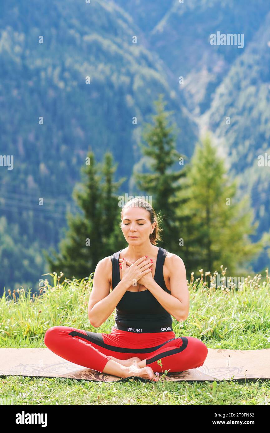 Outdoor portrait of healthy woman practicing yoga in mountains, sitting in lotus pose, holding hands on chest Stock Photo