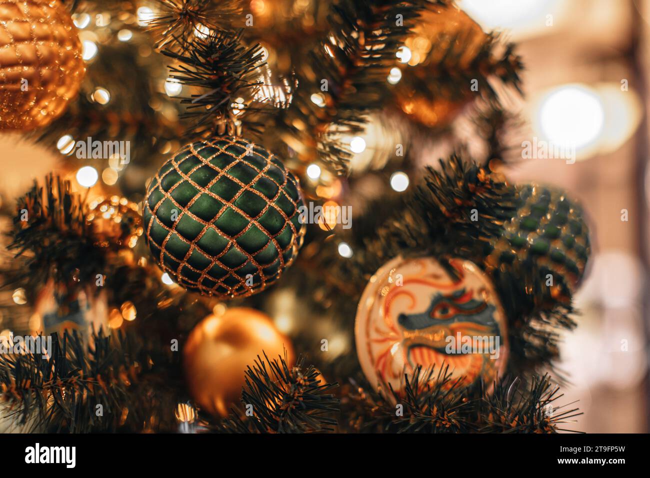Festive golden green shiny Christmas ball with a dragon pattern hanging on a Christmas tree with garland lights. Decorated spruce branches Stock Photo