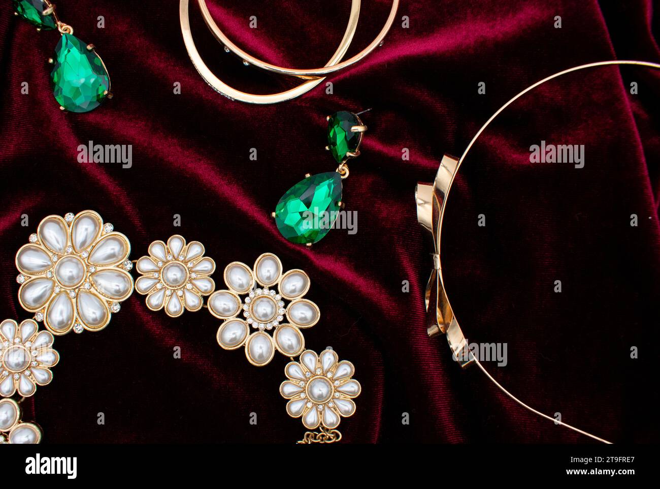Flat lay photo of a christmas party outfit with gold and green earrings, jewelry, and pearl necklace on a red velvet dress background Stock Photo