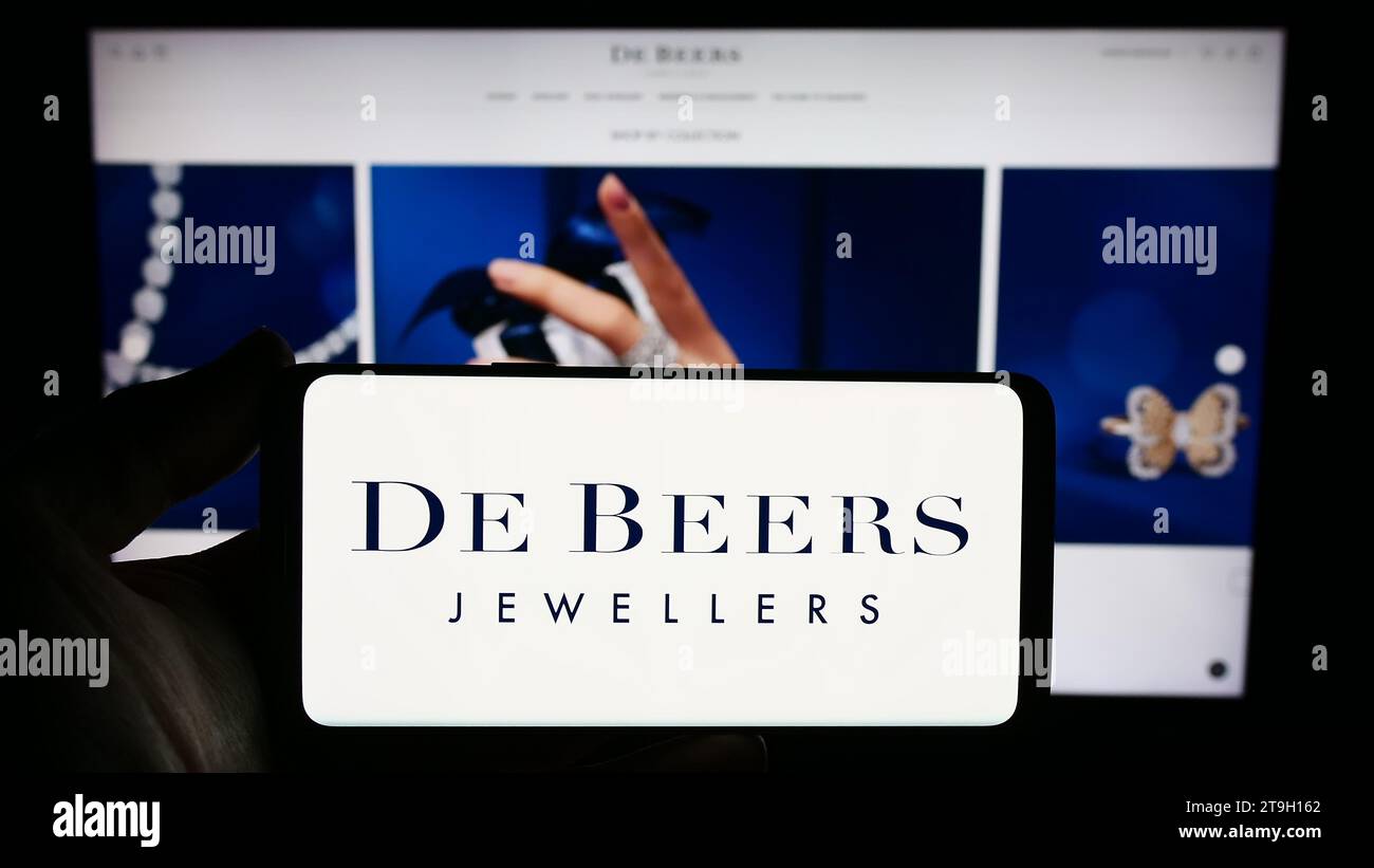Person holding smartphone with logo of diamond mining and trading company De Beers Group in front of website. Focus on phone display. Stock Photo