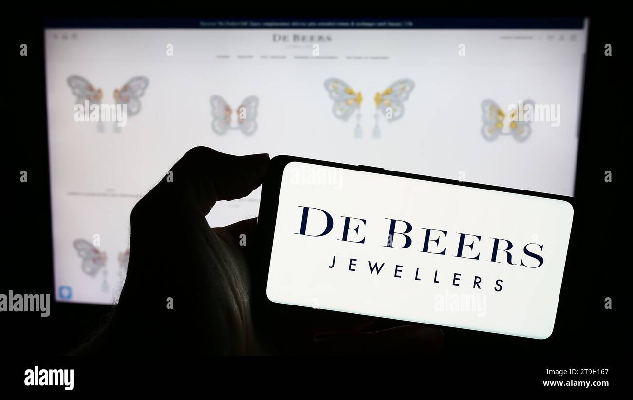 Person holding mobile phone with logo of diamond mining and trading company De Beers Group in front of business web page. Focus on phone display. Stock Photo