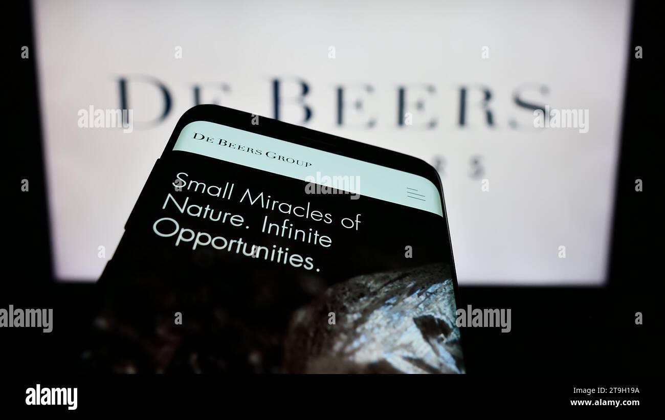 Mobile phone with webpage of diamond mining and trading company De Beers Group in front of business logo. Focus on top-left of phone display. Stock Photo