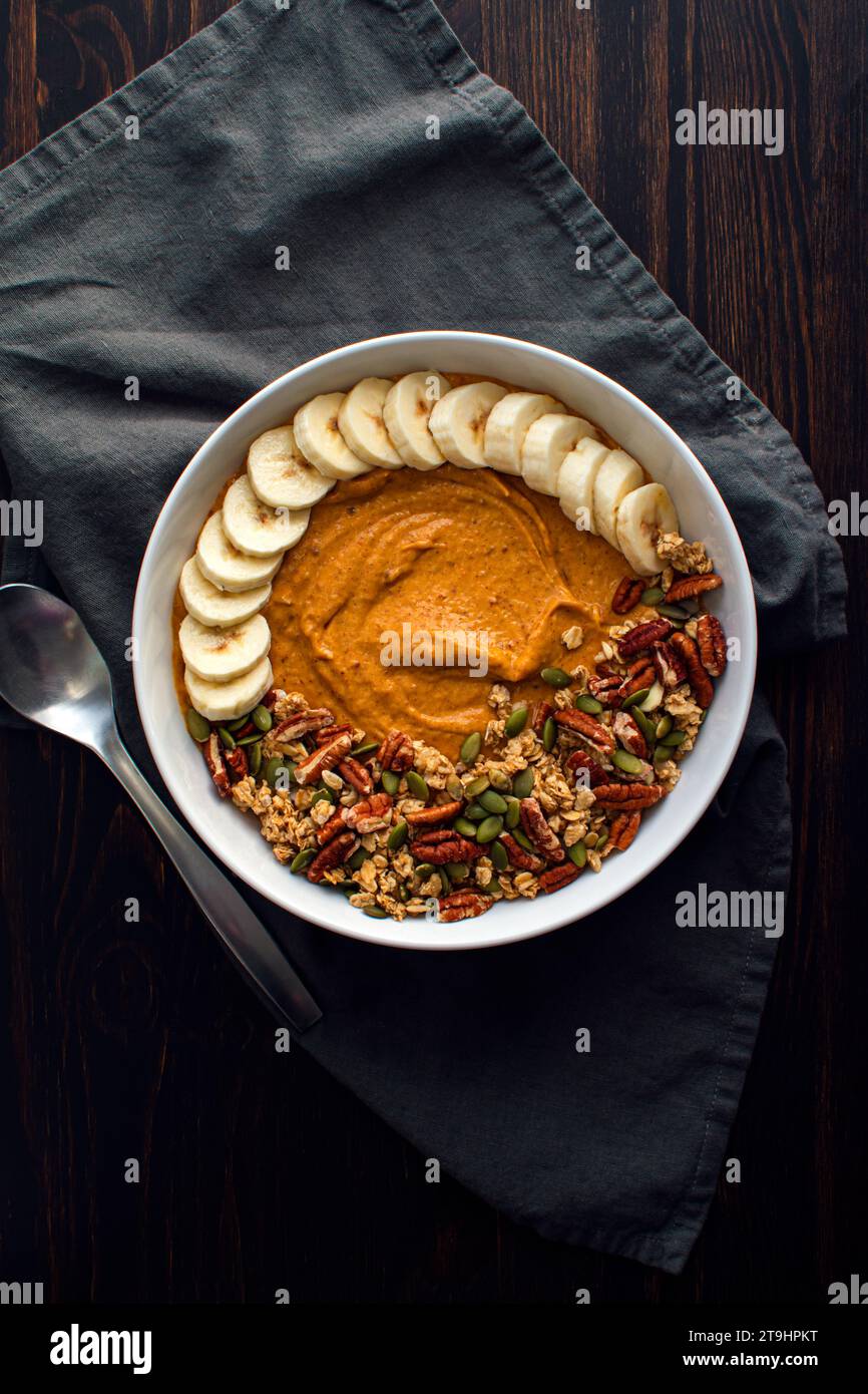 Pumpkin Pie Smoothie Bowl on a Wooden Table with a Spoon: Pumpkin pie smoothie topped with granola, pecans, pumpkin seeds, and sliced bananas Stock Photo