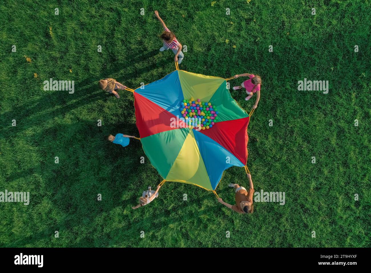 Group of children and teachers playing with rainbow playground parachute on green grass, top