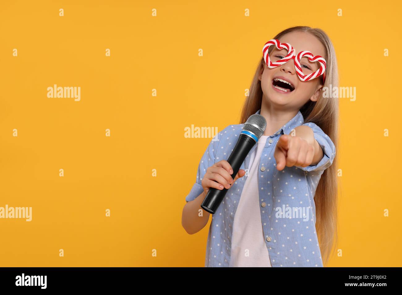 Cute little girl with funny glasses and microphone singing on yellow background, space for text Stock Photo
