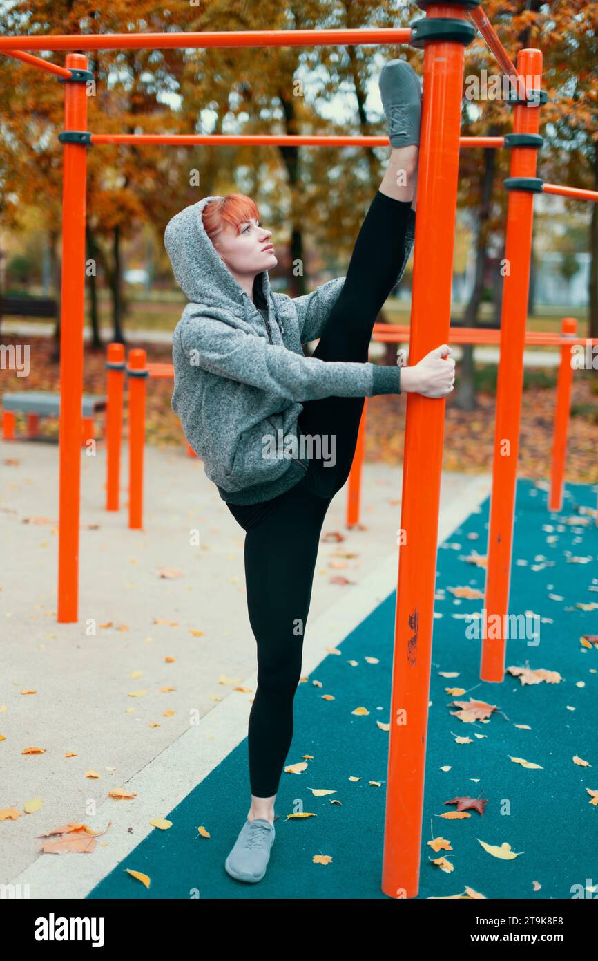 Young hooded Caucasian gymanst woman warming up at playground pillars and doing splits in park at autumn Stock Photo
