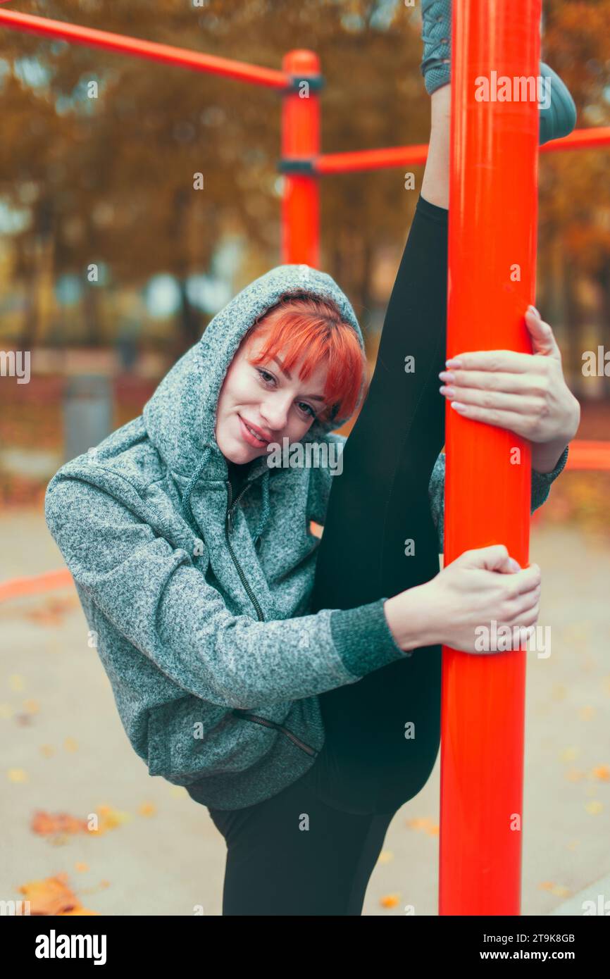 Young hooded parkour woman warming up at playground pillars and doing splits in park at autumn Stock Photo