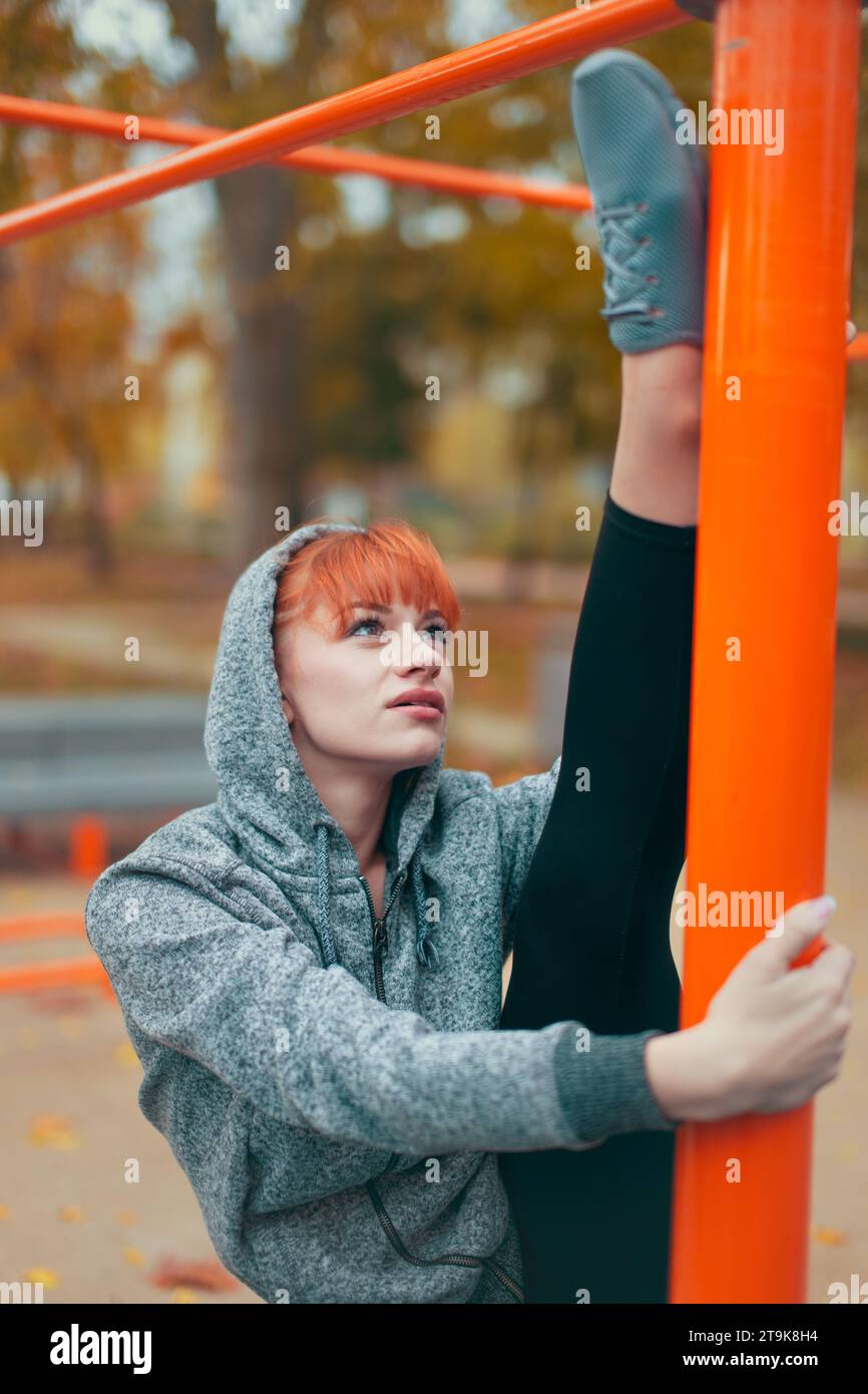 Young hooded Caucasian woman warming up at playground pillars and doing splits at autumn in park Stock Photo