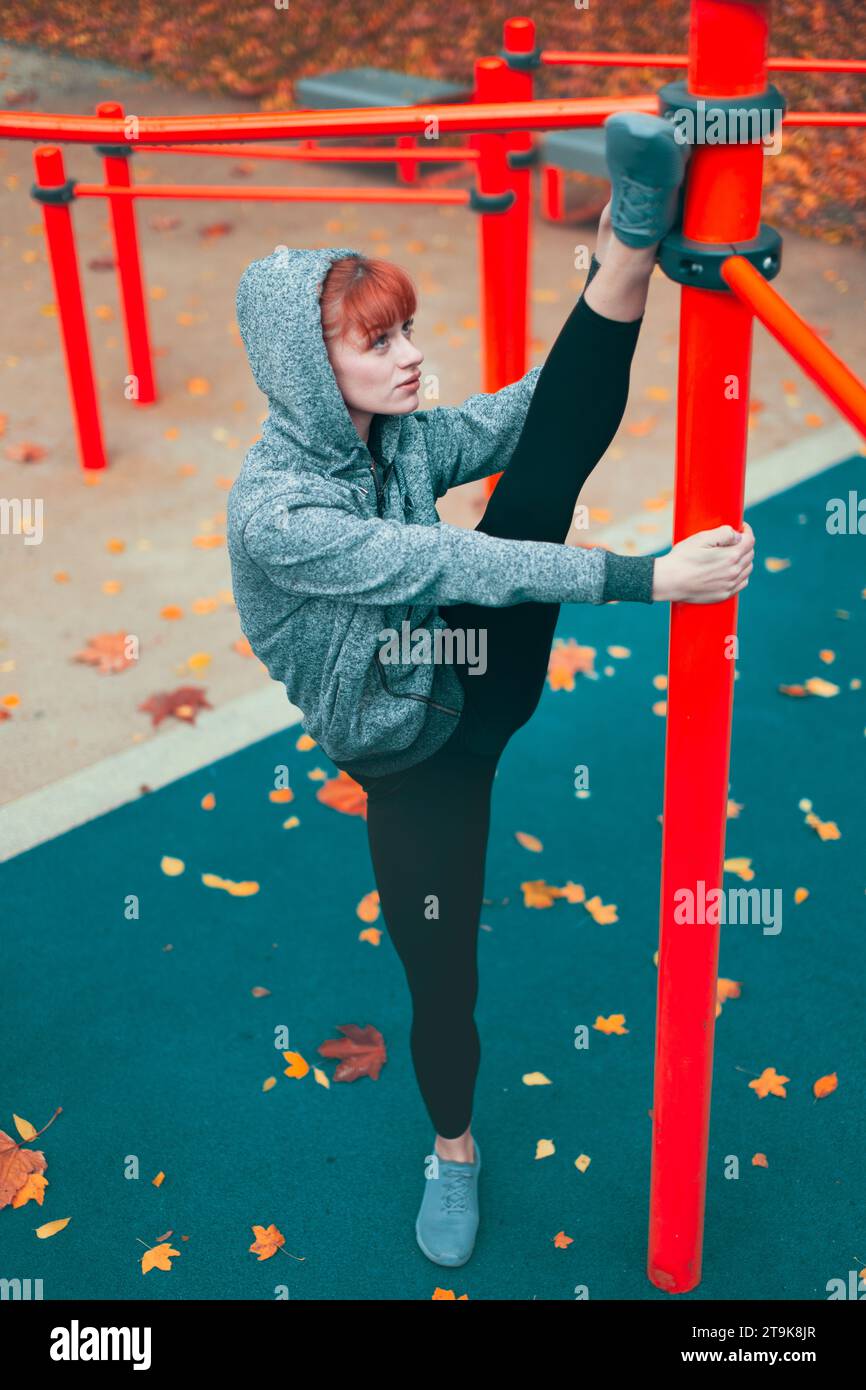 Young hooded Caucasian gymanst woman warming up at playground pillars and doing splits Stock Photo