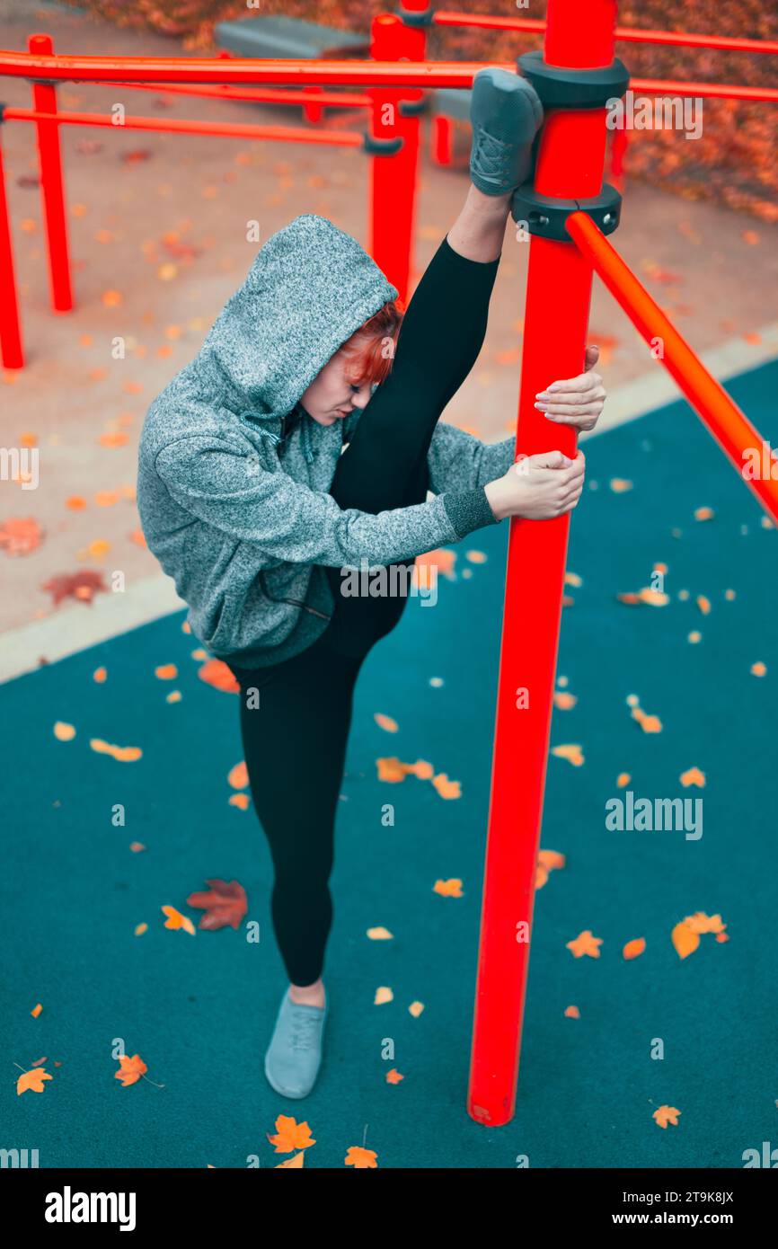 Young hooded Caucasian martial artist woman warming up at playground pillars and doing splits Stock Photo