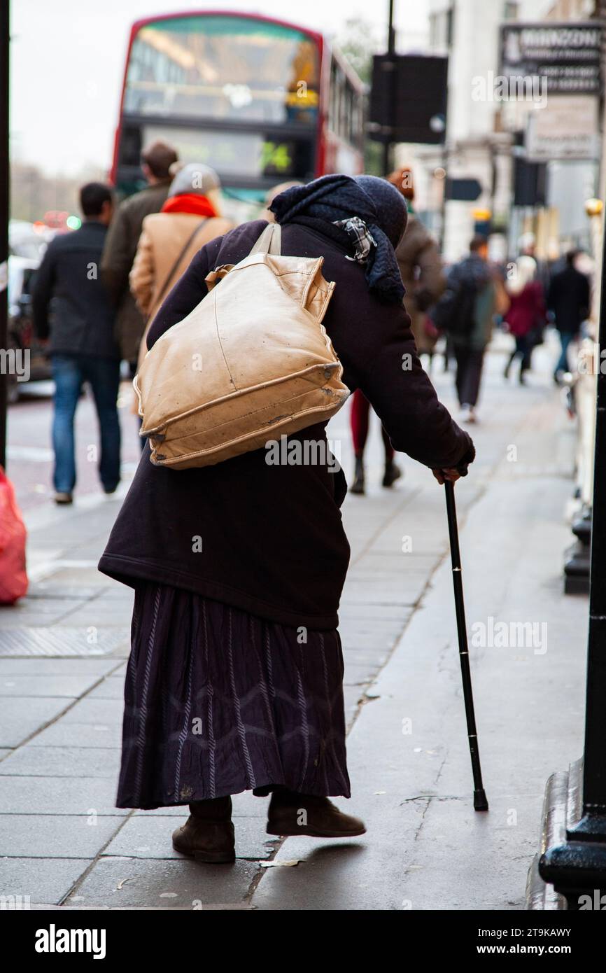 back view of elderly stooped lady using walking stick and wearing decaying bag, taking a step along a busy central London high street Stock Photo