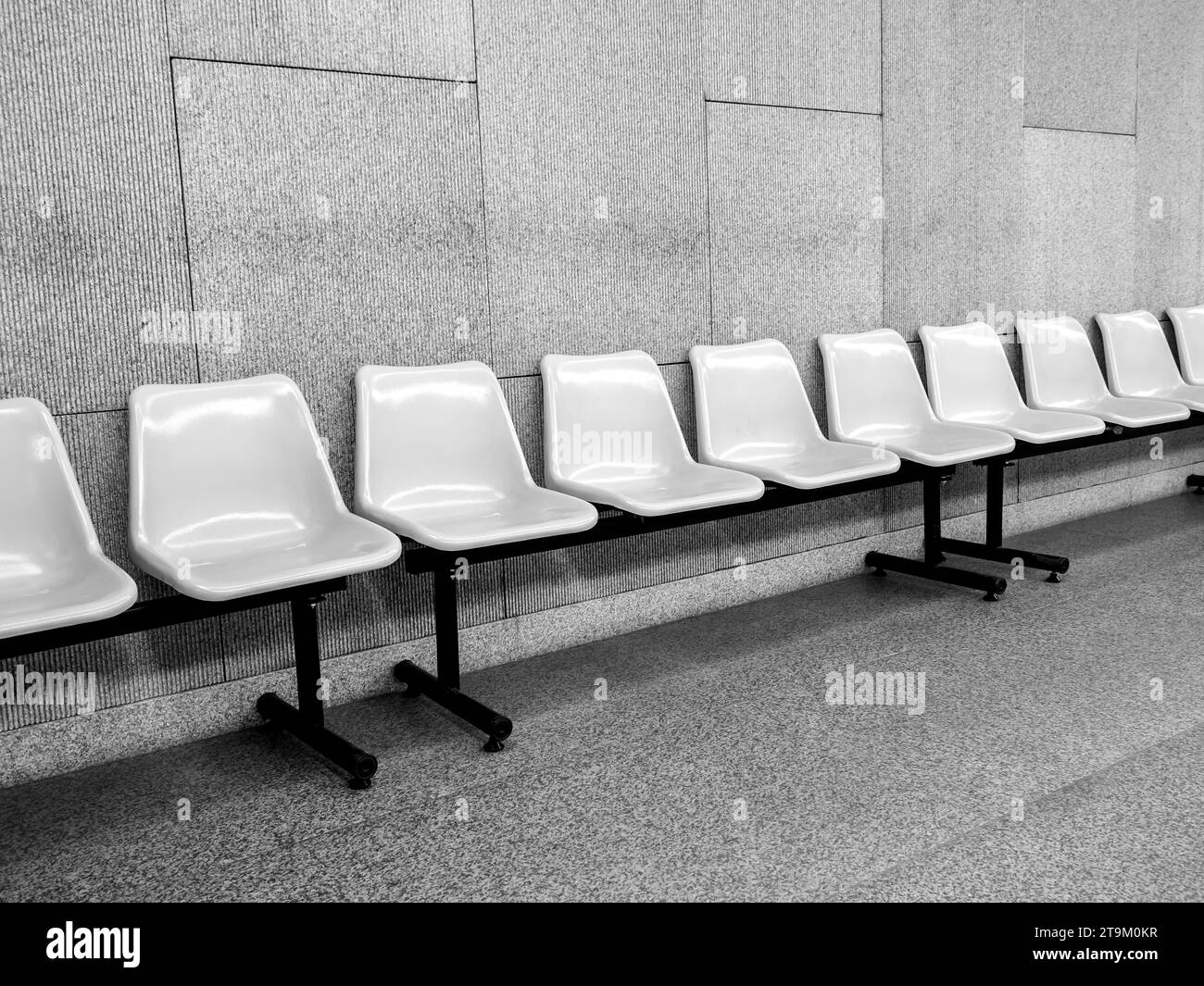 Black and white photo scene of row of empty waiting chair with white plastic seats with black iron legs seats on grey tiles and concrete wall backgrou Stock Photo