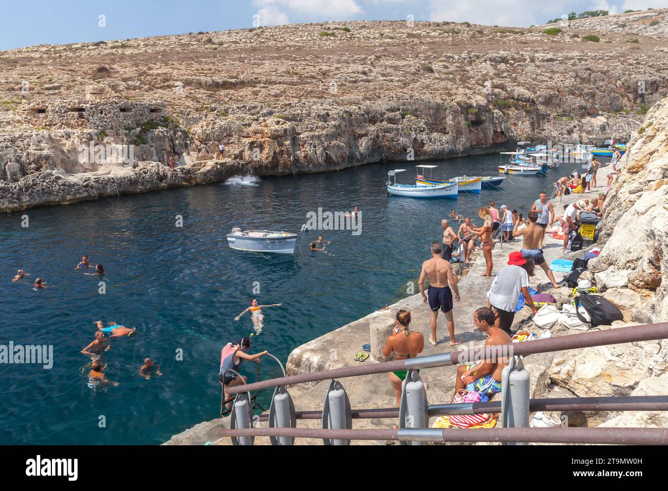 Blue Grotto, Malta - August 22, 2019: Coastal view with the Blue Grotto harbor. Wooden boats are anchored in front of public beach full of people on a Stock Photo