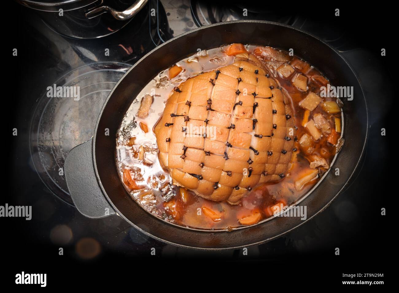 Roast pork with rind in diamond shapes and larded with cloves in a casserole with vegetables and broth on a black stove top, cooking a festive meal, h Stock Photo