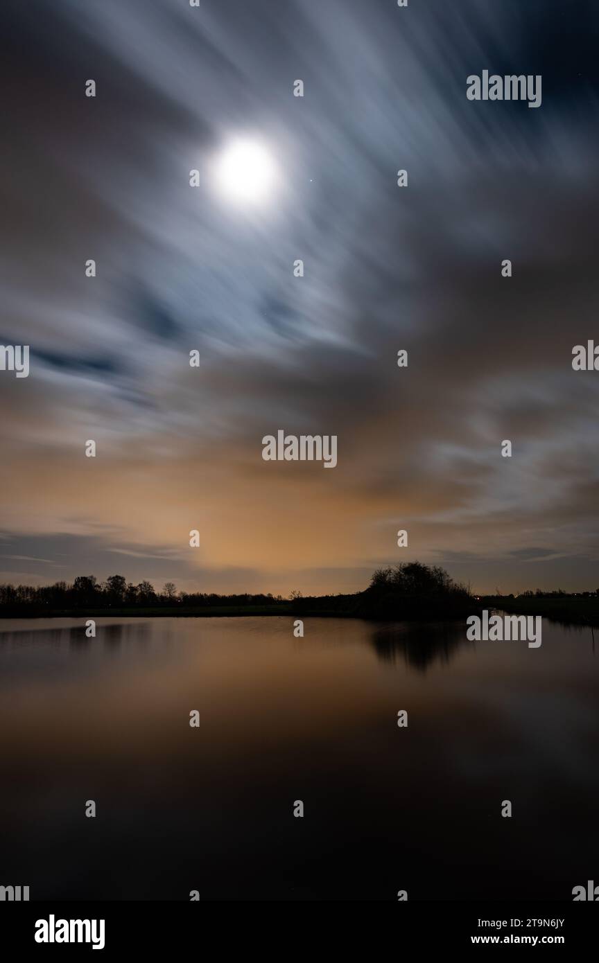 Scenic nighttime image of the moon over a lake while clouds pass by. Stock Photo