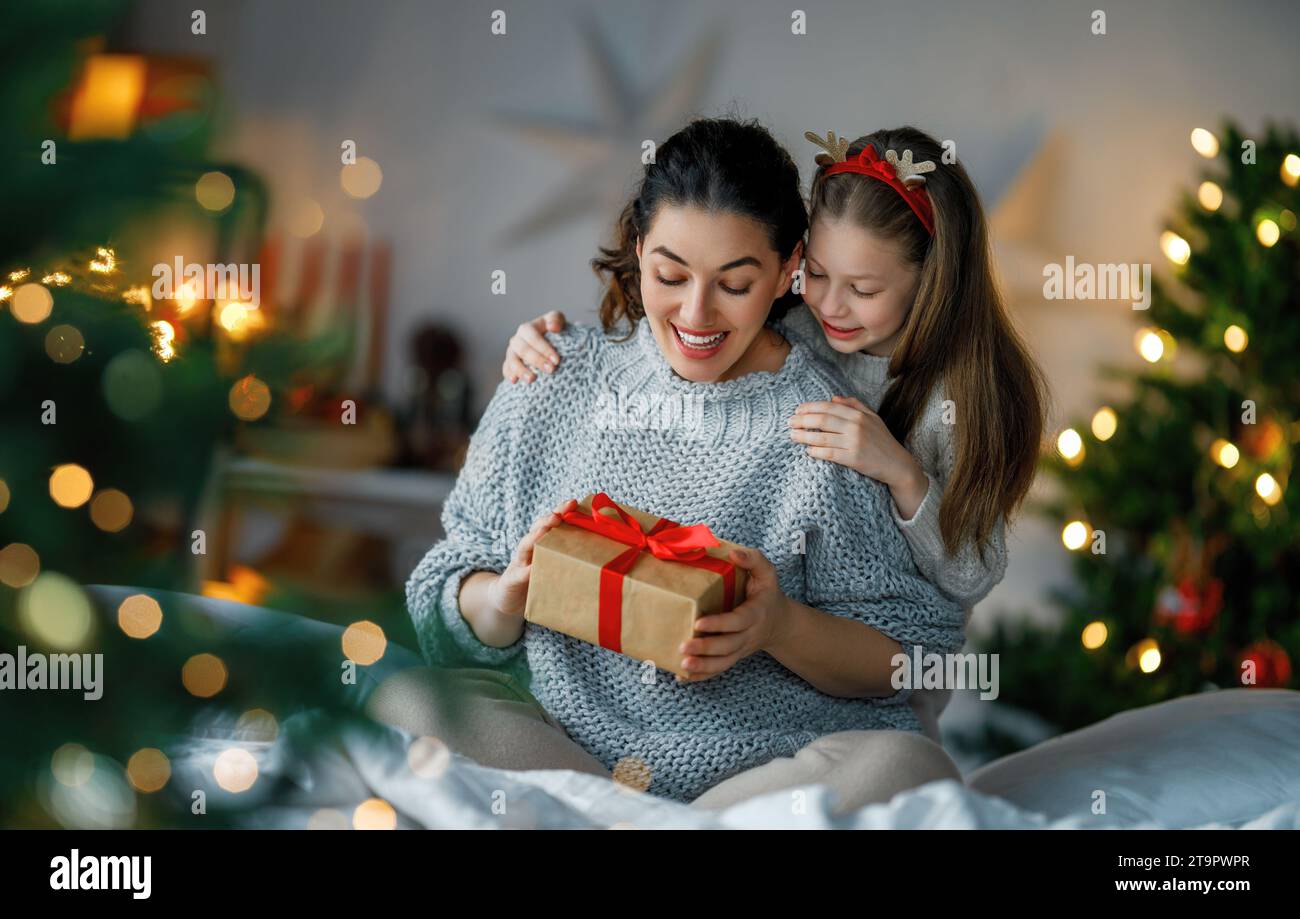 Merry Christmas and Happy Holidays. Cheerful mom and her cute daughter girl exchanging gifts. Parent and little child having fun near tree indoors. Lo Stock Photo