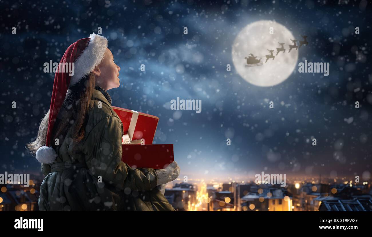 Merry Christmas. Cute little child with xmas present. Santa Claus flying in his sleigh against moon sky. Happy kid enjoy the holiday. Portrait of girl Stock Photo