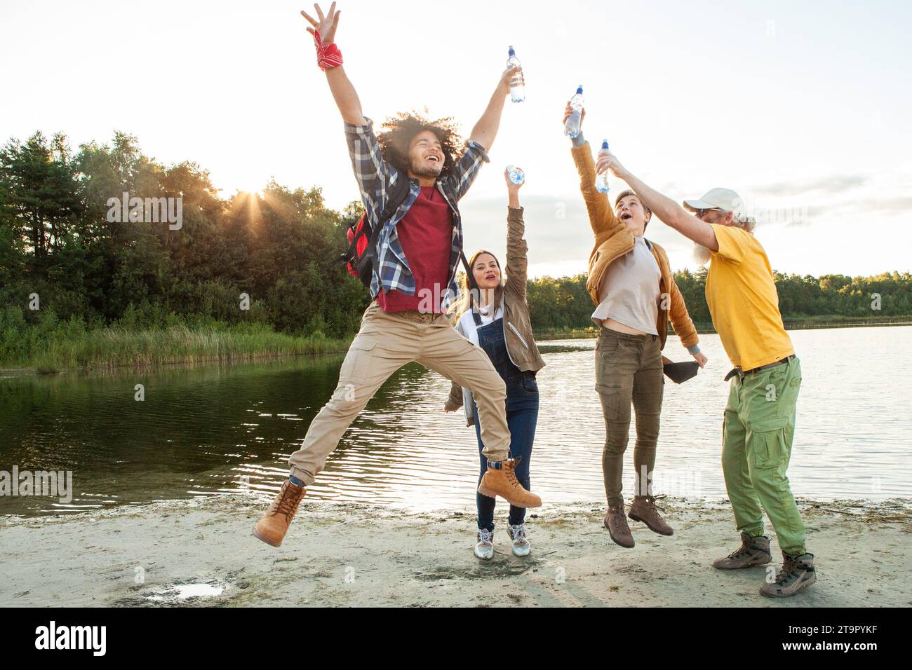 Capturing the essence of unbridled joy, this image features a group of friends in mid-celebration by the lake, one leaping high with arms and legs outstretched. Their faces are alive with laughter and excitement, each person caught in a moment of spontaneous delight. The setting sun casts a soft, golden light on the scene, reflecting off the water and adding a sense of warmth and vivacity to the carefree spirit of the gathering. Exuberant Joy by the Lakeside. High quality photo Stock Photo