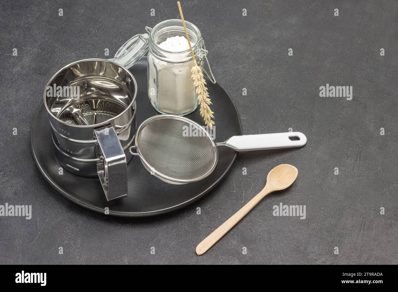 Mug for sifting flour and small sieve, glass jar with flour on black