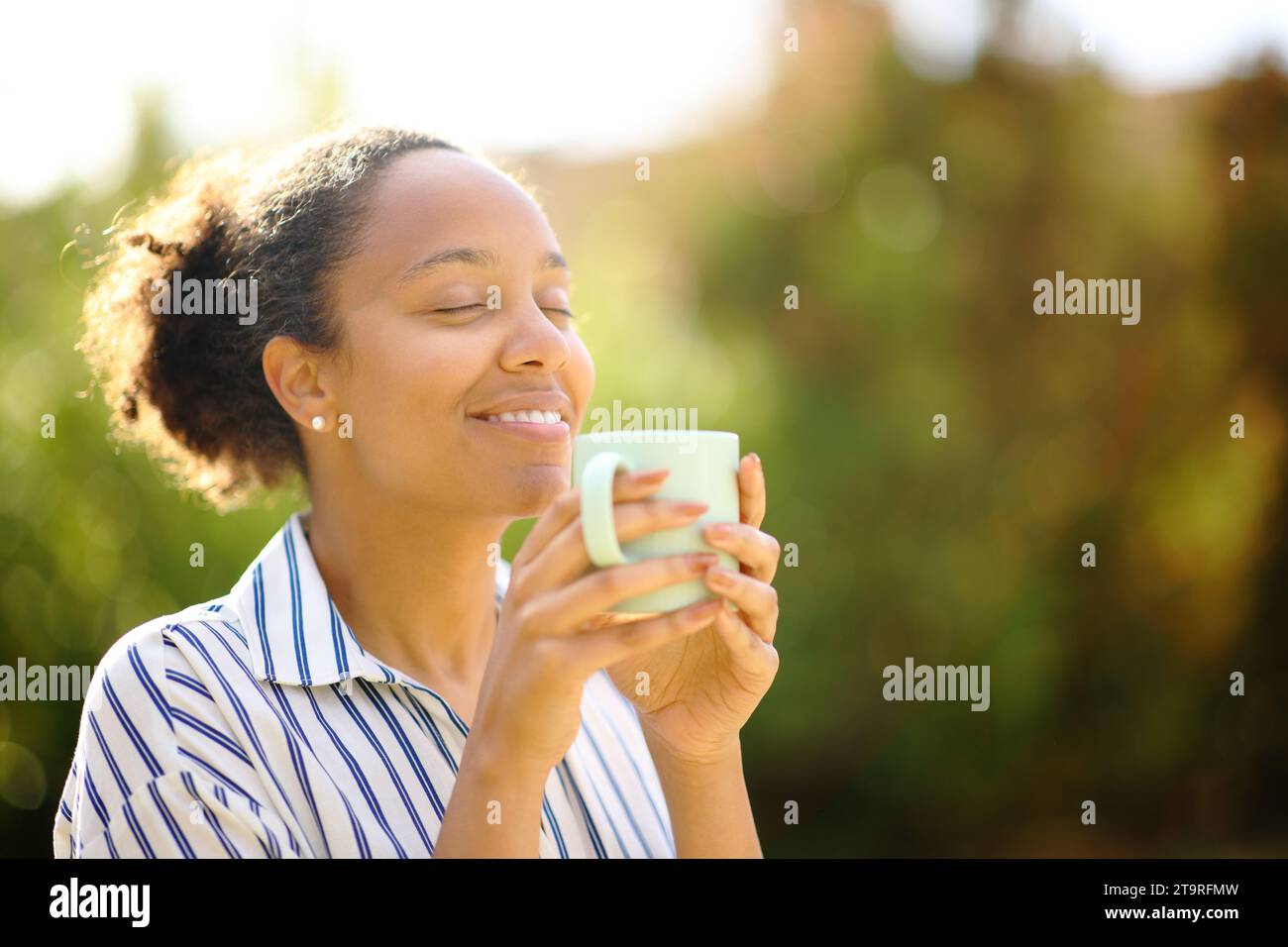 Happy black woman is smelling coffee standing in a park Stock Photo