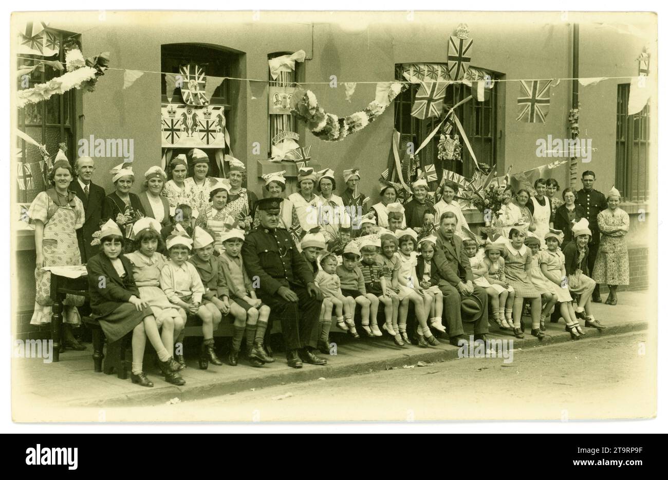 Original 1930's era postcard, charming image of smiling young children wearing party hats, posing for a photo in their street with their parents / adults, before a celebratory tea party, to celebrate the Coronation of George V1 (late Queen Elizabeth's father) Lots of bunting, flags, banners and party hats and characters. Fashions of the time. Two South Wales policemen attending to make sure they all don't get too excited. Eleanor Street, Tonypandy, Rhondda, south Wales, U.K. 1937. Stock Photo