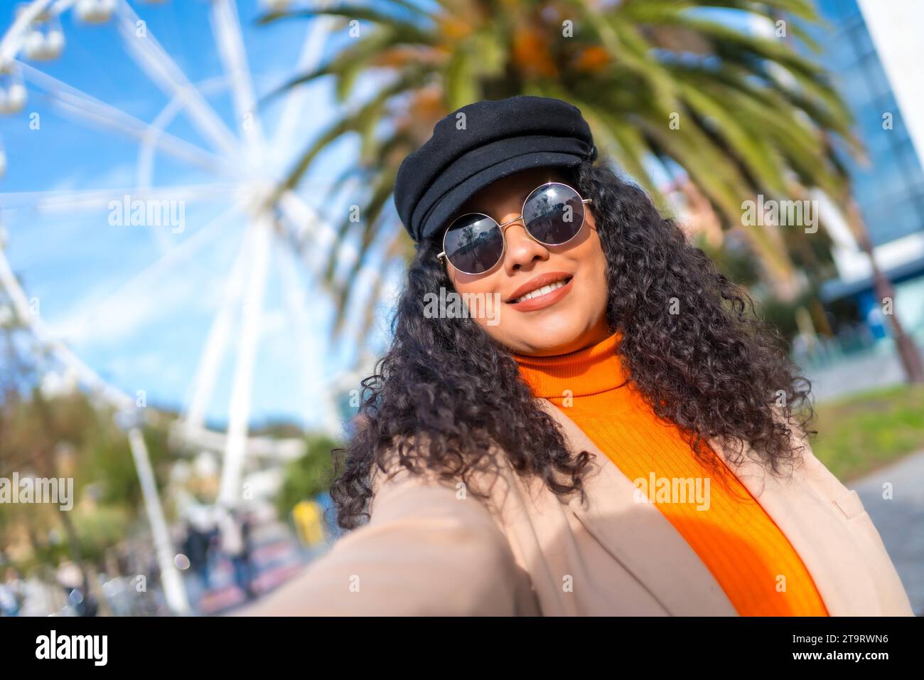 Beauty latin woman with sunglasses and hat taking a selfie in the city next to a ferris wheel in a sunny day of winter Stock Photo