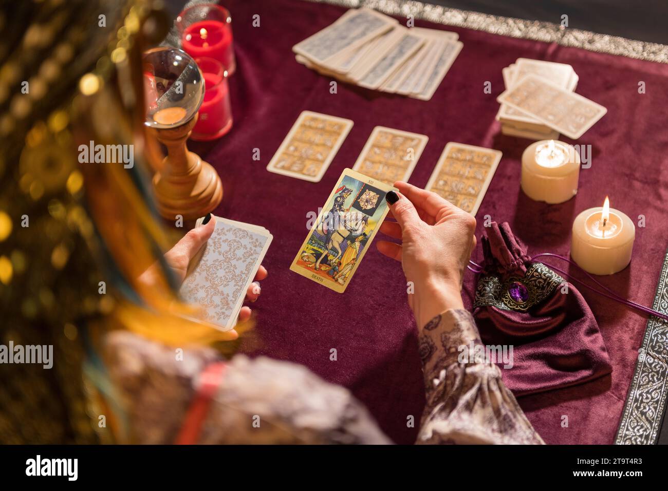 Back view of crop unrecognizable female fortune teller in costume picking death tarot card while sitting at table with burning candles and crystal Stock Photo