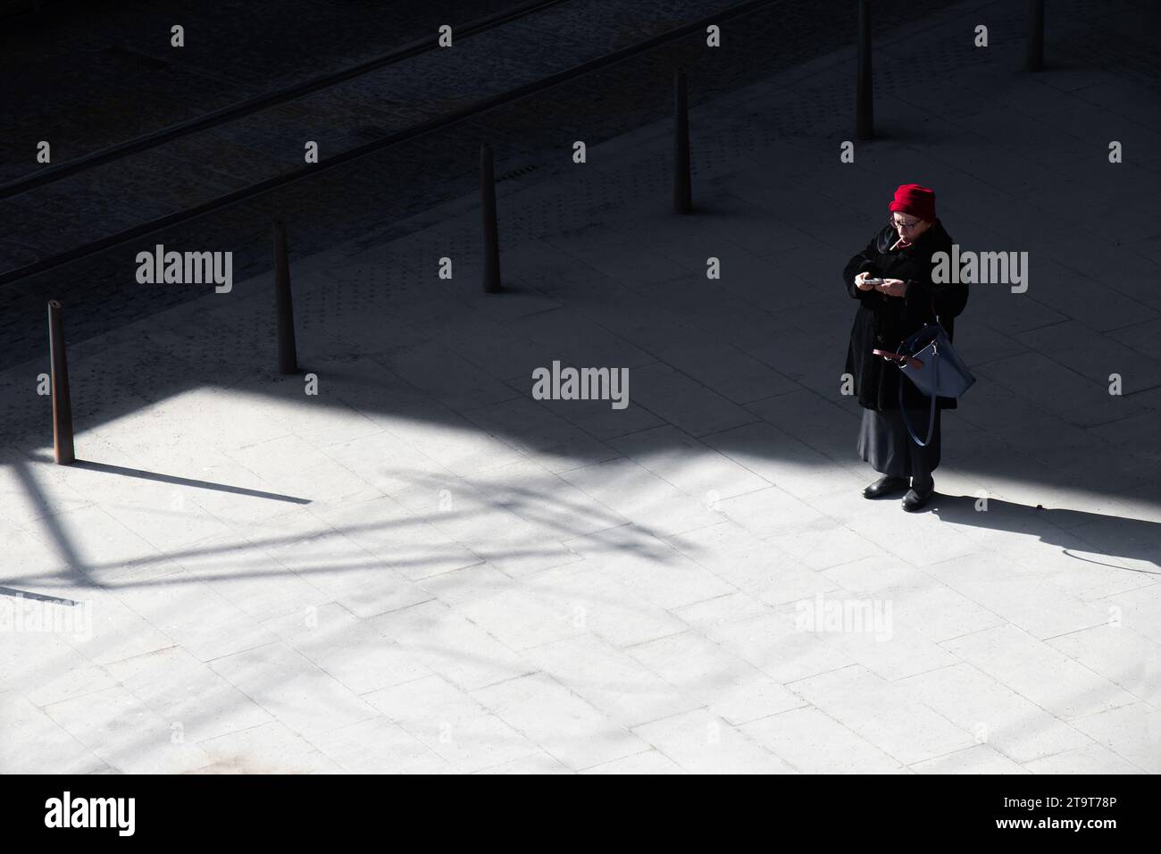 Belgrade, Serbia- March 18, 2022: One elderly woman with red hat standing alone on a town square street on the cold sunny day, high angle view Stock Photo