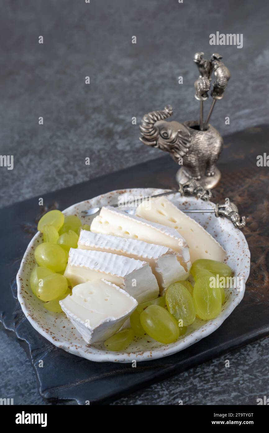 Camembert Brie cheese with grapes in a handmade clay craft plate on a dark background. Side view. Stock Photo