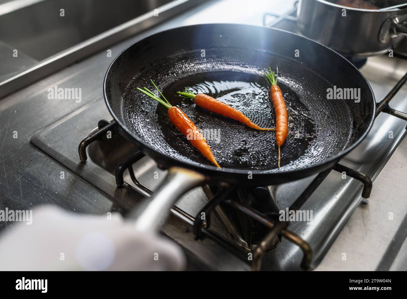 Hand tossing carrots in an oiled pan at a gas stove in a professional