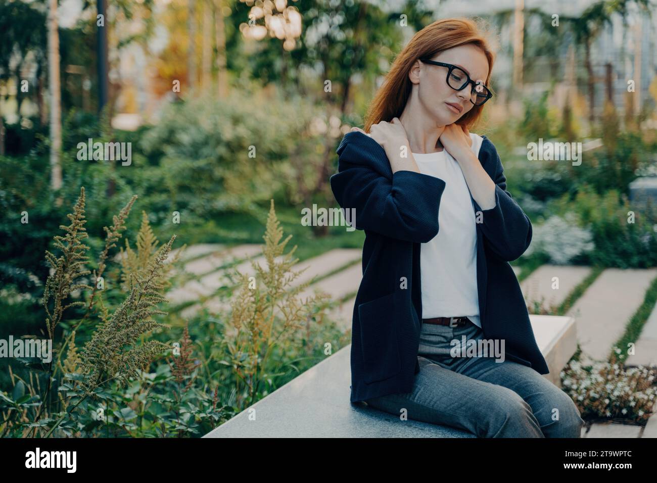 Businesswoman in a park taking a break from work, massaging her neck, amidst greenery Stock Photo