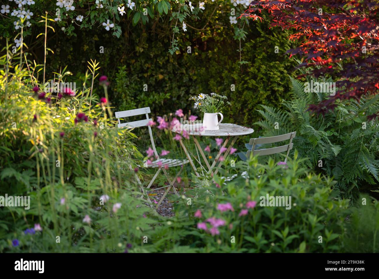 secluded garden seating area in the shade with metal cafte style table and chairs and jug of flowers - UK Stock Photo