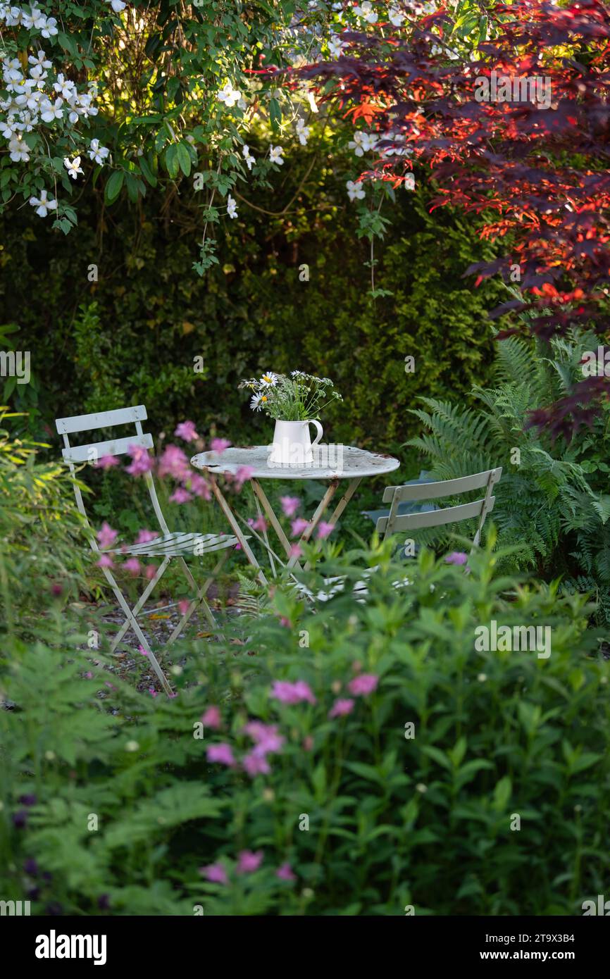 secluded garden seating area in the shade with metal cafte style table and chairs and jug of flowers - UK Stock Photo
