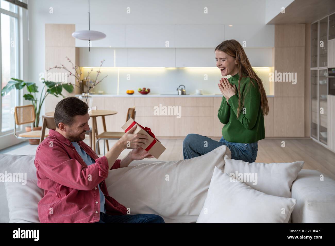 Laughing woman sitting on sofa, looking with excitement as beloved man trying to unwrap giftbox Stock Photo