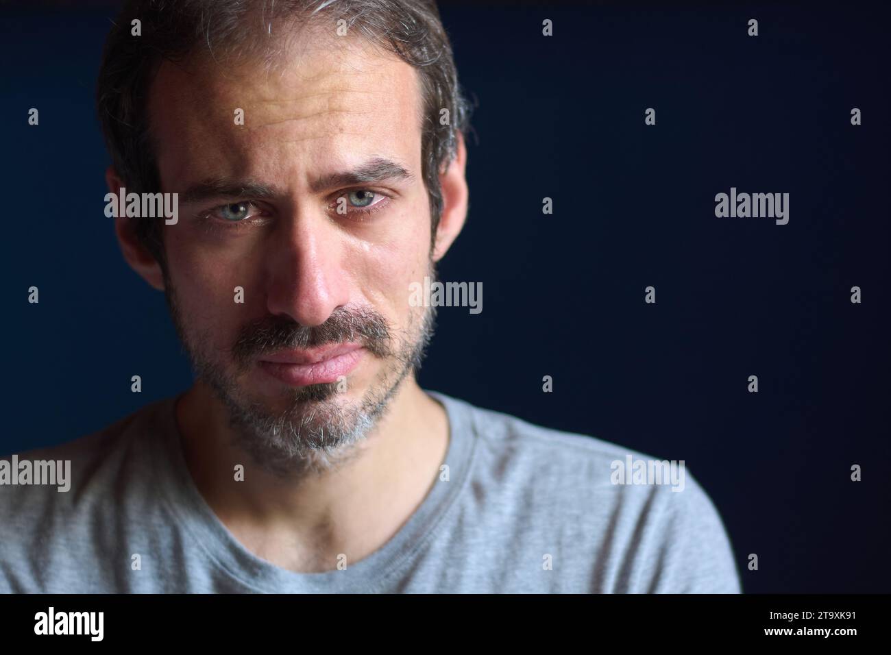 Close up of young caucasian 30 year old man with short beard and blue eyes crying while looking at the camera. Concept of depression, loss and grief. Stock Photo