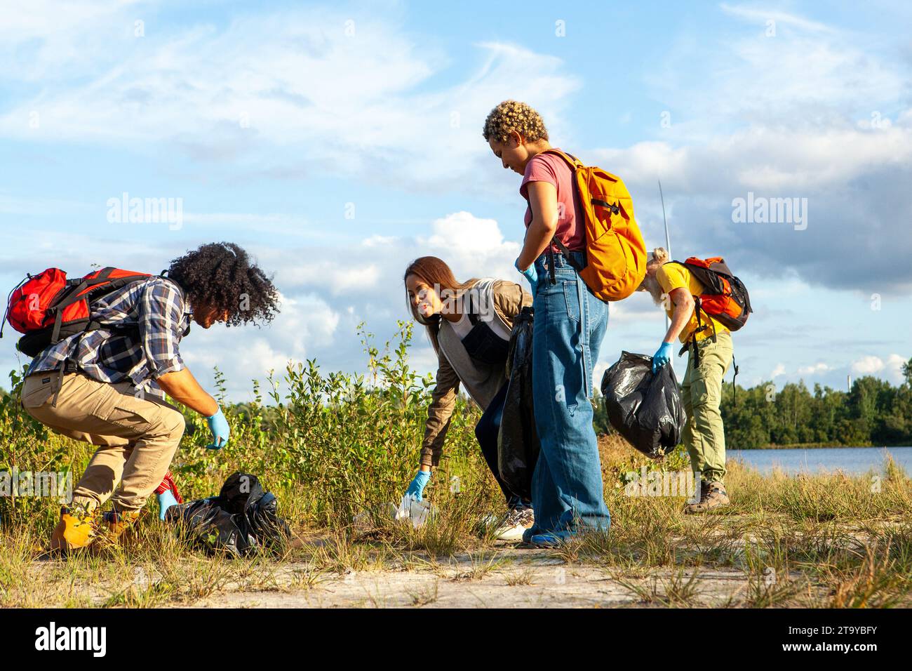 The photograph showcases a diligent group of volunteers engaged in a cleanup activity, demonstrating their commitment to environmental health. Each individual is equipped with gloves and bags, attentively removing litter from the natural landscape. Their focused actions under the open sky, near the water, reflect a collective dedication to preserving the beauty and integrity of the outdoor environment for future generations. Dedication to the Environment: Group Cleanup Action. High quality photo Stock Photo