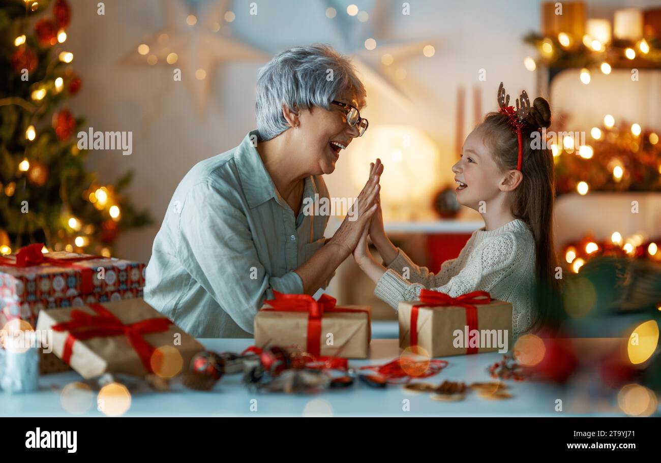 Happy Holidays. Cheerful grandmother and her cute granddaughter girl preparing for Christmas. People wrapping gifts, decorating home. Loving family wi Stock Photo