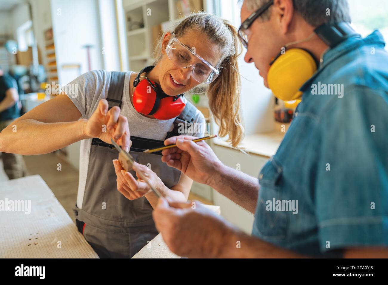 Carpentry mentor and apprentice measuring a wooden piece in a well-lit workshop Stock Photo