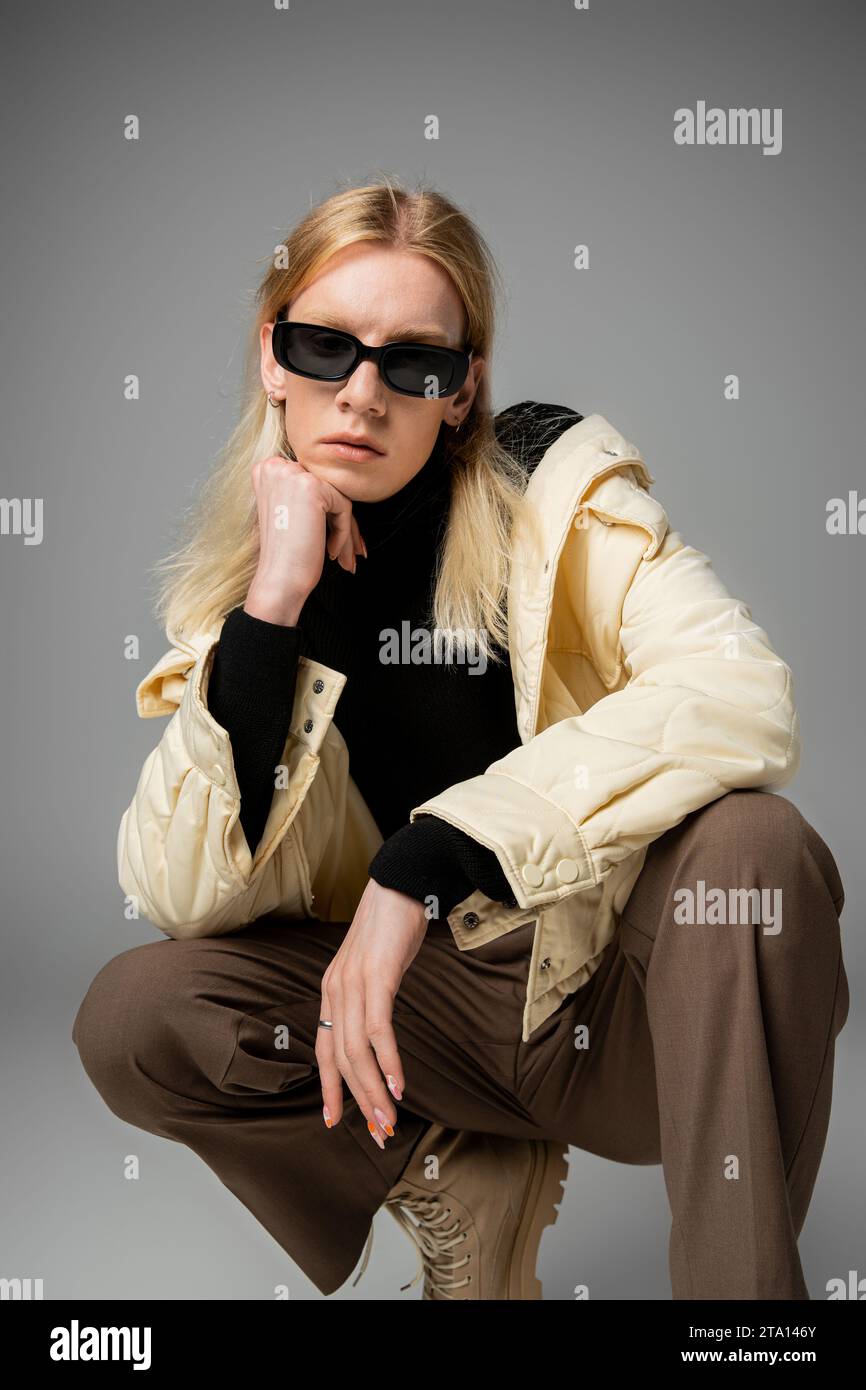 appealing androgynous person in stylish outfit with sunglasses squatting with fist under chin Stock Photo