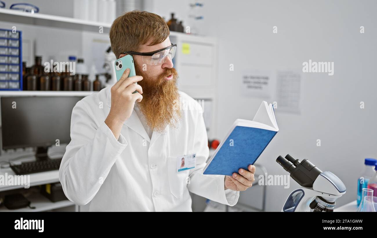 Chatty young redhead guy, a scientist engrossed in reading a book, takes a quick break indoors his laboratory to talk on his smartphone Stock Photo
