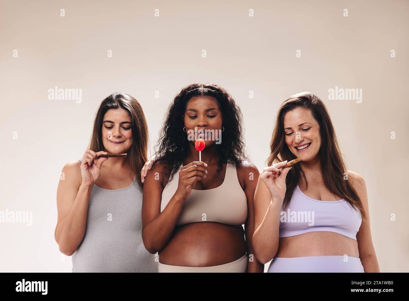 Young, diverse pregnant women in the third trimester stand together in a  studio. They wear jeans and bras, showcasing their baby bumps. Hugging and  looking at the camera, they represent a supportive