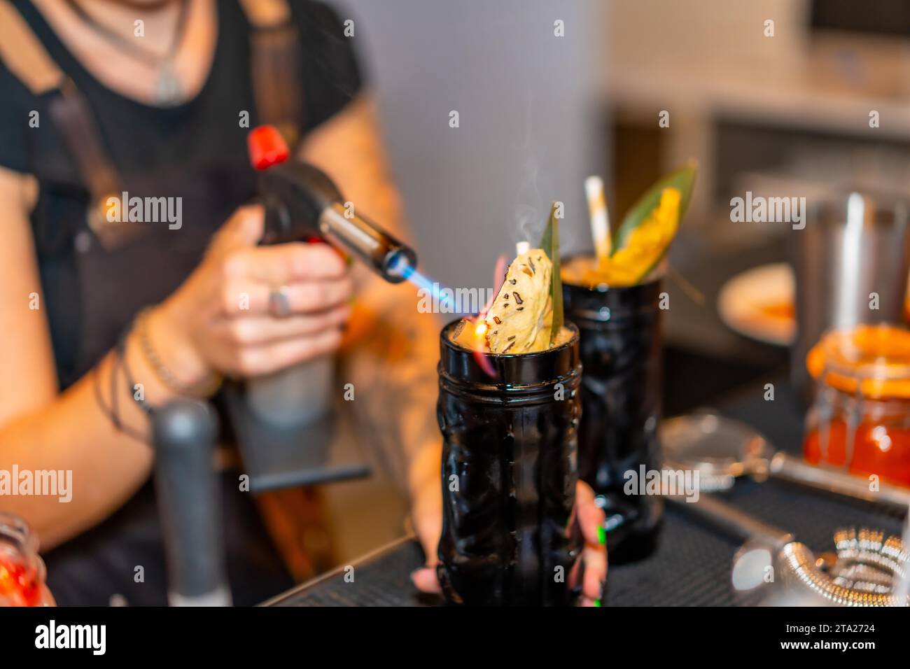Cropped photo with close-up of a female bartender flaming the garnish of a cocktail with a torch in the counter of a bar Stock Photo