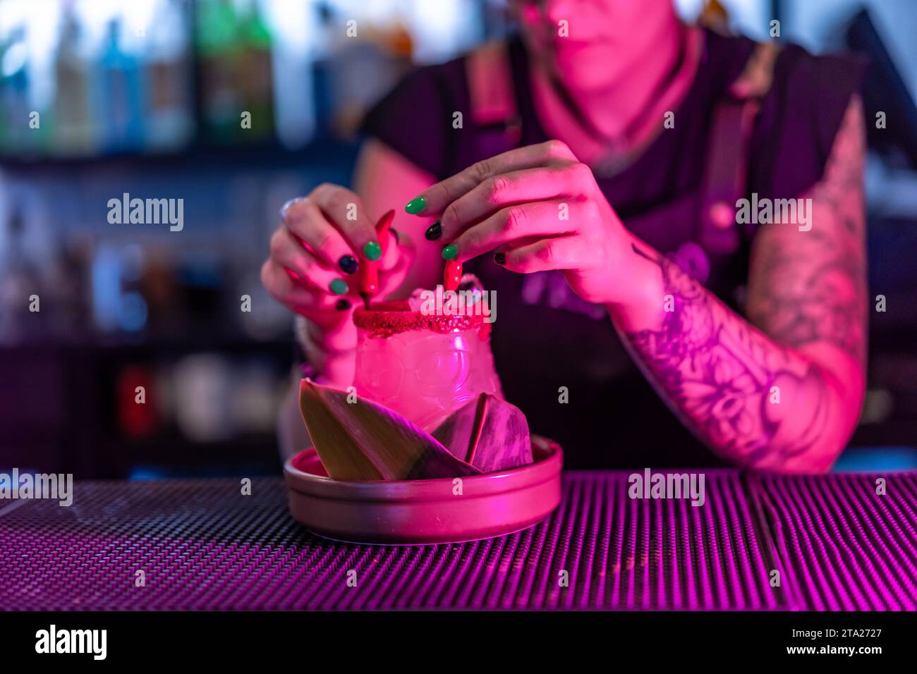 Close-up photo of a female alternative bartender decorating a margarita cocktail with devil horns in the counter of a night bar with neon lights Stock Photo