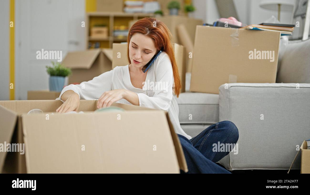 Young redhead woman unpacking cardboard box speaking on the phone at new home Stock Photo