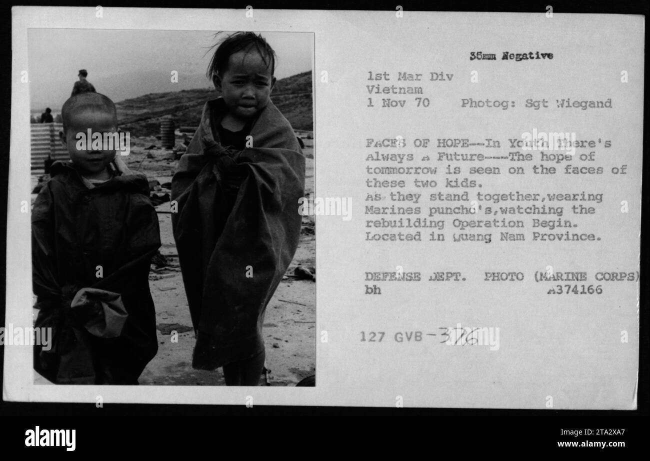 Two Vietnamese children, wearing Marine's ponchos, observe the
