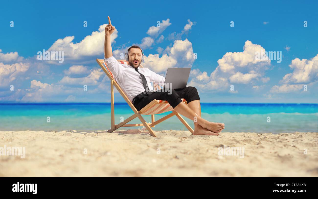 Cheerful businessman with headphones and laptop sitting on a beach ...