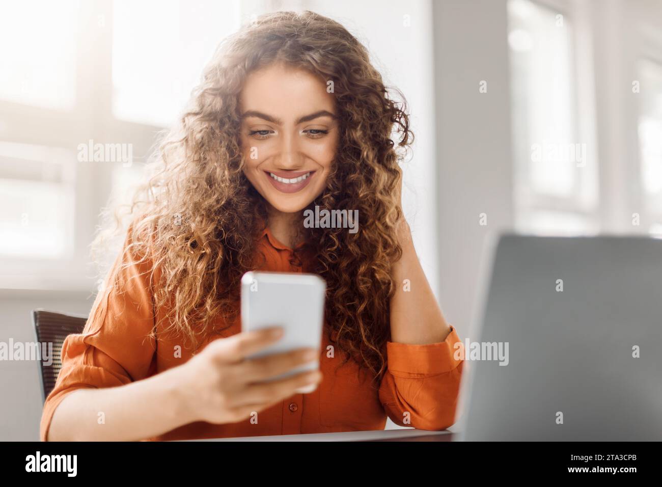 Happy curly-haired woman using smartphone at workplace Stock Photo