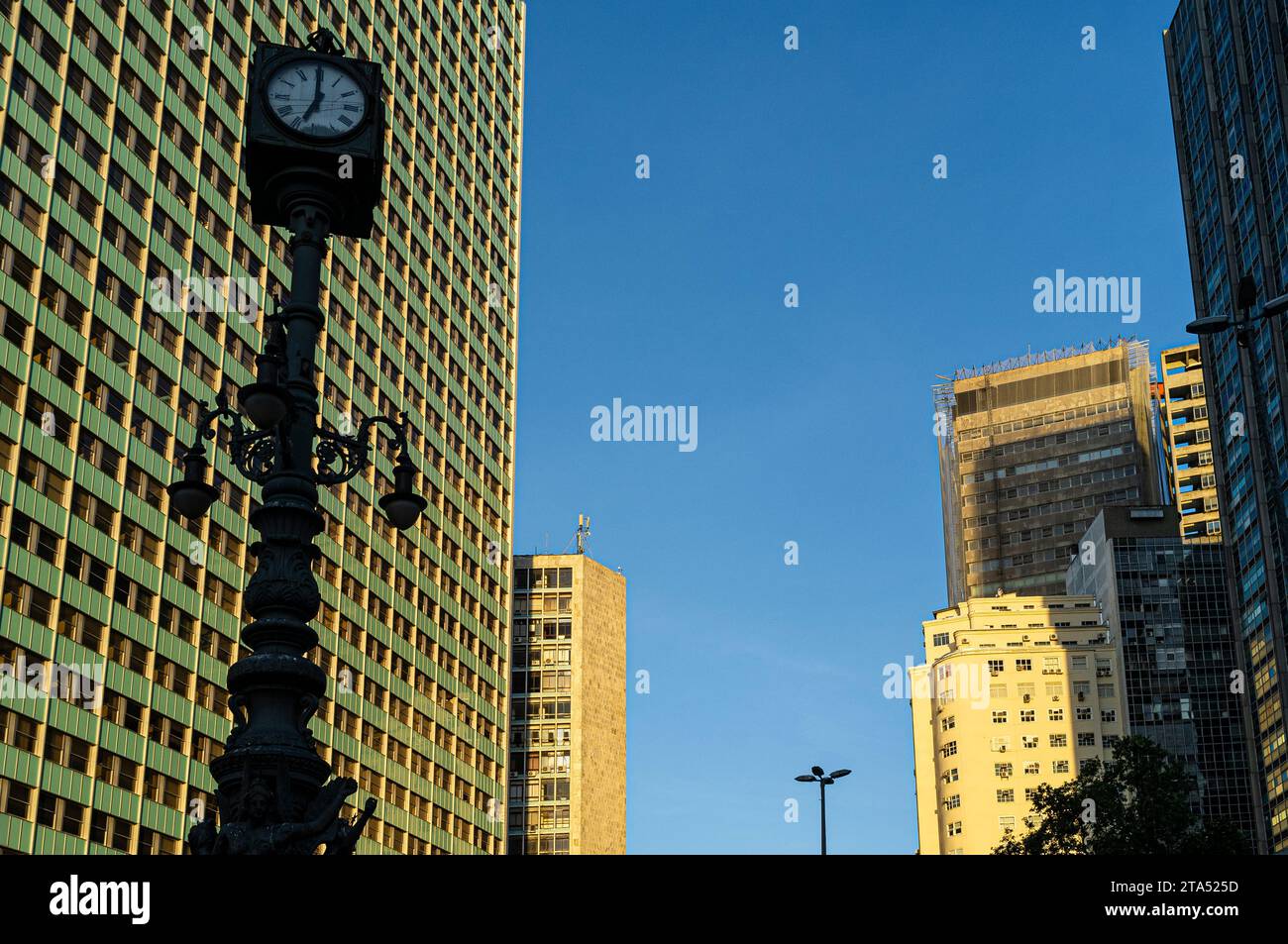 Largo da Carioca Clock in downtown Rio de Janeiro, surrounded by corporate buildings. Made in 1909 by the Brazilian Iron and Bronze Foundry Kobler and Co., it is equipped with an iron lamp holder, topped by a four-faced clock, and decorated by three allegorical figures of winged mermaids, representing, commerce, industry and navigation. Harmonious coexistence between ancient and modern - lamppost from the Belle Époque period share the urban space with corporate buildings. Stock Photo