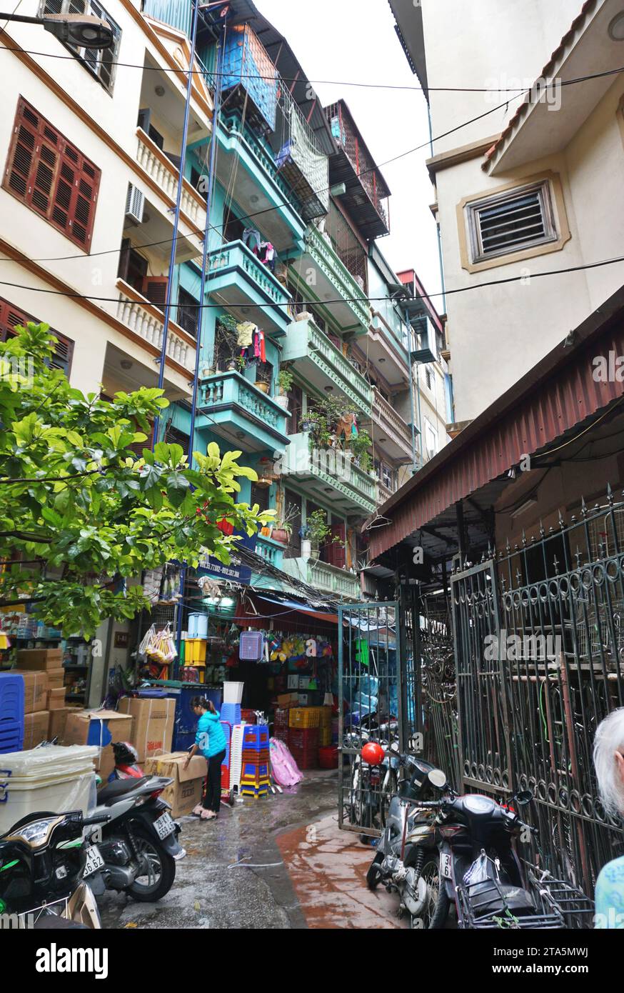 Multi-story apartment buildings line a narrow street in Hanoi, Vietnam. Shopfronts fill the ground level with motorcycle parking on the sidewalks Stock Photo